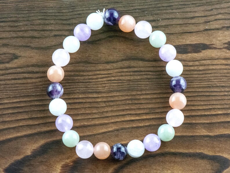 May include: A beaded bracelet featuring a variety of colorful gemstones. The bracelet includes round beads in shades of purple, peach, white, and green. The bracelet is displayed on a dark wooden surface.