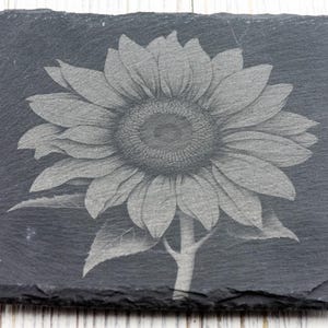 K&ouml;nnte beinhalten: Ein schwarzer Schieferuntersetzer mit einem lasergravierten Design einer Sonnenblume. Die Sonnenblume ist in Graut&ouml;nen und Wei&szlig; dargestellt.