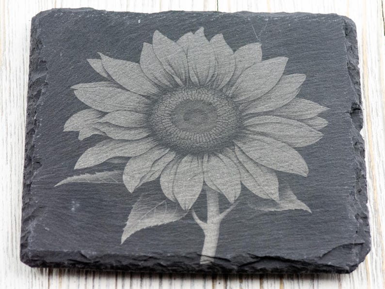 K&ouml;nnte beinhalten: Ein schwarzer Schieferuntersetzer mit einem lasergravierten Design einer Sonnenblume. Die Sonnenblume ist in Graut&ouml;nen und Wei&szlig; dargestellt.