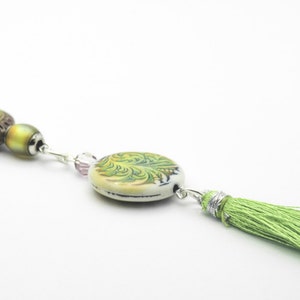 May include: A green and brown beaded necklace with a green tassel. The necklace features a round, white ceramic bead with a green floral design.
