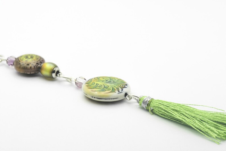 May include: A green and brown beaded necklace with a green tassel. The necklace features a round, white ceramic bead with a green floral design.