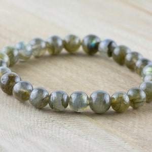 Puede incluir: Un brazalete de cuentas de labradorita pulida. Las cuentas redondas son principalmente de color gris verdoso con destellos iridiscentes azules y dorados. El brazalete es el&aacute;stico y descansa sobre una superficie de madera clara.