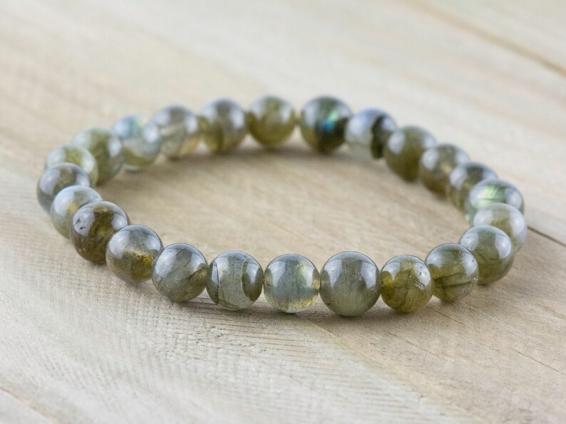 Puede incluir: Un brazalete de cuentas de labradorita pulida. Las cuentas redondas son principalmente de color gris verdoso con destellos iridiscentes azules y dorados. El brazalete es el&aacute;stico y descansa sobre una superficie de madera clara.