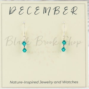 May include: Teal crystal dangle earrings on a silver hook. The earrings are displayed on a card that says "DECEMBER" and "Black Brook Shop". The card also says "Nature-Inspired Jewelry and Watches".