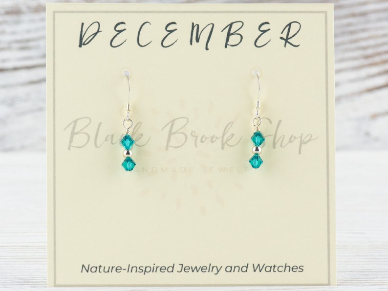 May include: Teal crystal dangle earrings on a silver hook. The earrings are displayed on a card that says "DECEMBER" and "Black Brook Shop". The card also says "Nature-Inspired Jewelry and Watches".