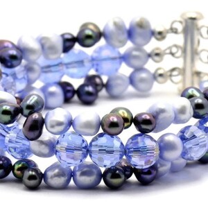 May include: A multi-strand bracelet featuring a mix of blue and black beads. The bracelet is made with glass and pearl beads, and has a silver clasp.