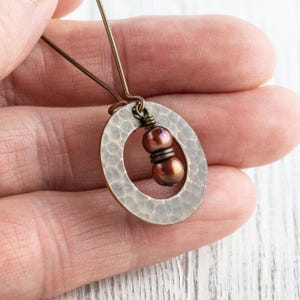 May include: A handcrafted, hammered metal pendant with an oval shape. The pendant features two brown beads and is suspended from a delicate wire. The jewelry is held in a hand, showcasing the intricate details and craftsmanship.