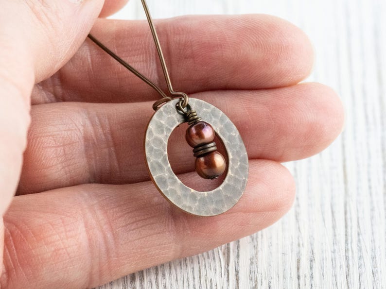May include: A handcrafted, hammered metal pendant with an oval shape. The pendant features two brown beads and is suspended from a delicate wire. The jewelry is held in a hand, showcasing the intricate details and craftsmanship.