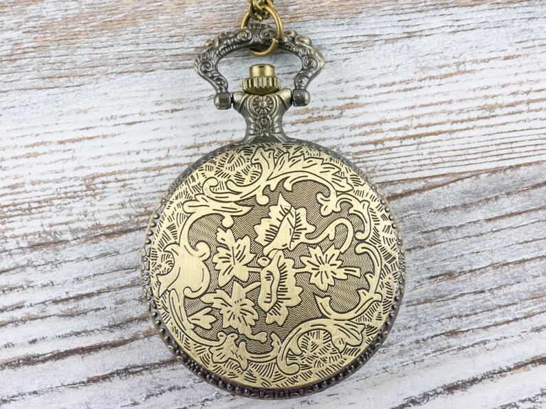 May include: A gold-toned pocket watch with a floral design on the front. The watch has a chain attached to the top.