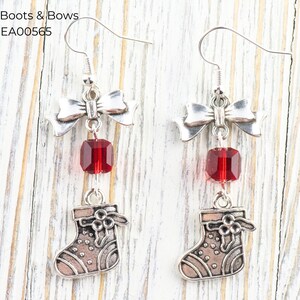 Pode incluir: Brincos pendurados em tom prateado com um la&ccedil;o, uma conta vermelha e um pequeno pingente de meia. Os brincos t&ecirc;m um tema festivo e de feriado. O texto "Red Boots & Bows" e "EA00565" &eacute; vis&iacute;vel.