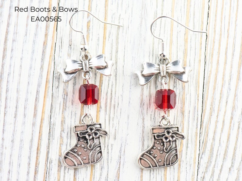 Pode incluir: Brincos pendurados em tom prateado com um la&ccedil;o, uma conta vermelha e um pequeno pingente de meia. Os brincos t&ecirc;m um tema festivo e de feriado. O texto "Red Boots & Bows" e "EA00565" &eacute; vis&iacute;vel.