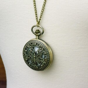 K&ouml;nnte beinhalten: Antike bronzefarbene Taschenuhr-Anh&auml;nger-Halskette. Das runde Uhrengeh&auml;use hat ein Schmetterlingsdesign mit floralen Akzenten. Die Uhr h&auml;ngt an einer passenden Kette.