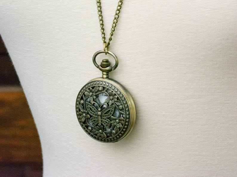 K&ouml;nnte beinhalten: Antike bronzefarbene Taschenuhr-Anh&auml;nger-Halskette. Das runde Uhrengeh&auml;use hat ein Schmetterlingsdesign mit floralen Akzenten. Die Uhr h&auml;ngt an einer passenden Kette.
