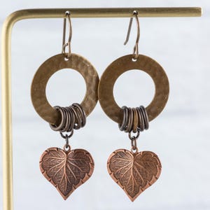 Op de afbeelding: Een paar handgemaakte oorbellen met gehamerde messing ringen en donkere metalen accenten. Elke oorbel heeft een koperkleurige hartvormige bedel met bladdetails, die onder de ring hangt. De oorbellen worden gepresenteerd op een goudkleurige standaard.