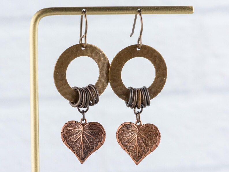 Op de afbeelding: Een paar handgemaakte oorbellen met gehamerde messing ringen en donkere metalen accenten. Elke oorbel heeft een koperkleurige hartvormige bedel met bladdetails, die onder de ring hangt. De oorbellen worden gepresenteerd op een goudkleurige standaard.