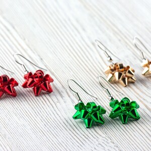 May include: Earrings in red, gold, and green bow shapes. The earrings have silver-colored hooks. The bows are shiny and appear to be made of a metallic material. The earrings are displayed on a white, wood-grain surface.