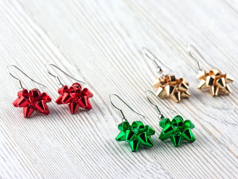 May include: Earrings in red, gold, and green bow shapes. The earrings have silver-colored hooks. The bows are shiny and appear to be made of a metallic material. The earrings are displayed on a white, wood-grain surface.