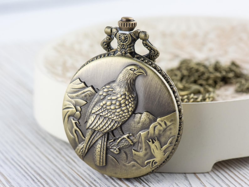 May include: A vintage-style pocket watch with a bronze finish. The watch face features an engraved image of a bird with detailed feathers and a mountain scene.
