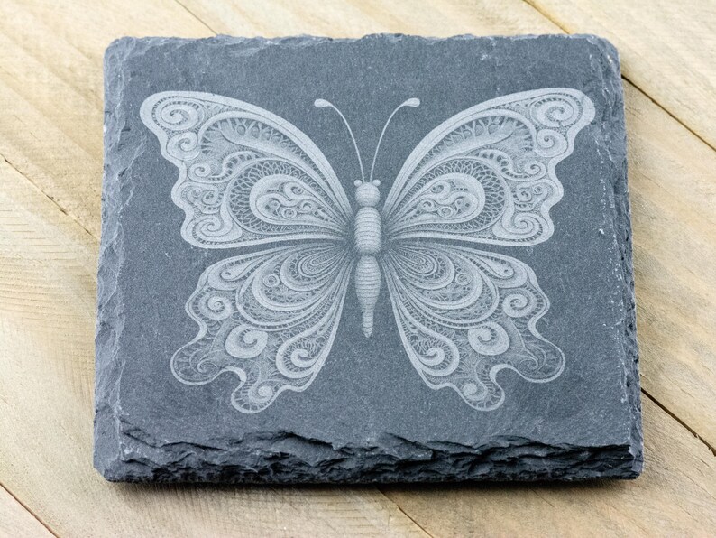 May include: A black slate coaster featuring a white, intricately designed butterfly etched into the surface.