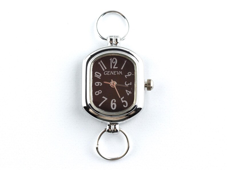 May include: Silver-toned Geneva watch with a rectangular face and a dark brown dial. The watch features white numerals and hands, with a small crown on the side. Includes two key rings for attachment.