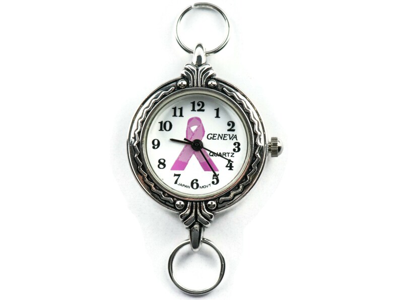 K&ouml;nnte beinhalten: Eine silberne Taschenuhr mit wei&szlig;em Zifferblatt und einem rosafarbenen Schleifendesign. Das Zifferblatt der Uhr zeigt "GENEVA QUARTZ" und hat schwarze Zeiger. Die Uhr hat eine silberne Kette mit einem Ring oben.