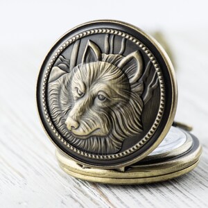 May include: A gold-toned pocket watch with a wolf's head design on the front. The wolf's head is detailed and has a textured fur pattern. The watch is open and the back is visible.