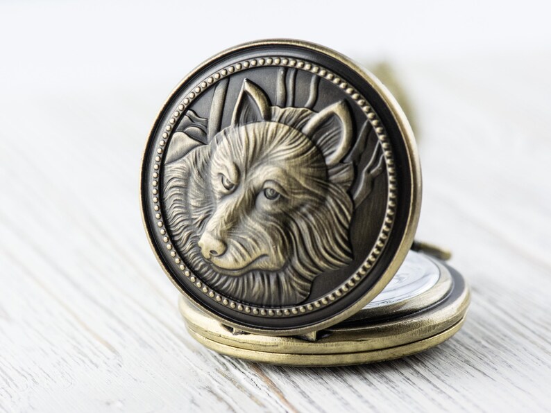 May include: A gold-toned pocket watch with a wolf's head design on the front. The wolf's head is detailed and has a textured fur pattern. The watch is open and the back is visible.