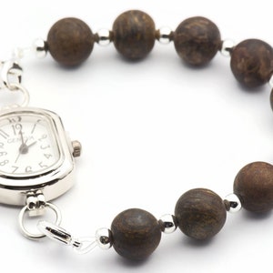 Puede incluir: Un reloj plateado con una esfera cuadrada y cuentas de madera marr&oacute;n unidas a la correa. La esfera del reloj tiene marcas de n&uacute;meros romanos negros y la palabra "GENOVA" impresa en ella.