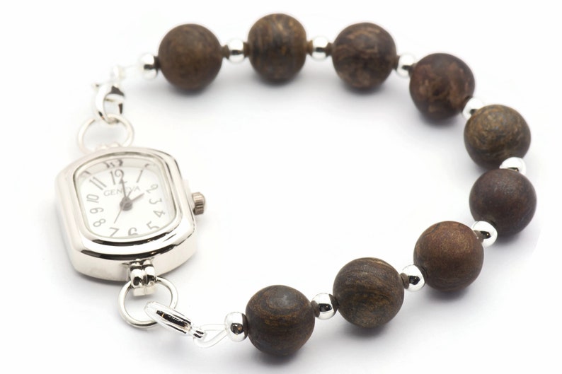Puede incluir: Un reloj plateado con una esfera cuadrada y cuentas de madera marr&oacute;n unidas a la correa. La esfera del reloj tiene marcas de n&uacute;meros romanos negros y la palabra "GENOVA" impresa en ella.