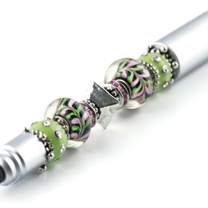 K&ouml;nnte beinhalten: Ein silberner Stift mit schwarzer Spitze und gr&uuml;nen und rosa Glasperlen. Die Perlen sind mit silbernen Akzenten und Strasssteinen verziert.