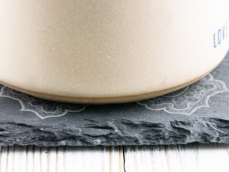 Puede incluir: Un taz&oacute;n de cer&aacute;mica beige con la palabra "LOVE" impresa en el lateral, sentado sobre un posavasos de pizarra negra con un dise&ntilde;o decorativo.