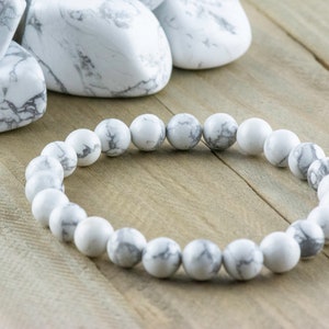 Pode incluir: Um bracelete de contas de howlite branco com veios cinzentos. O bracelete &eacute; feito de contas redondas e est&aacute; sobre uma superf&iacute;cie de madeira.