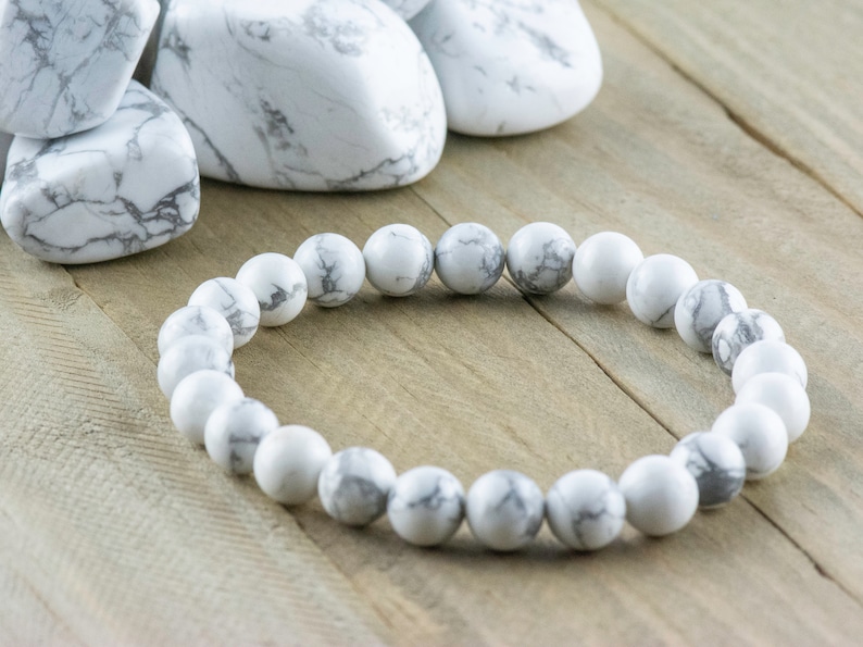 Pode incluir: Um bracelete de contas de howlite branco com veios cinzentos. O bracelete &eacute; feito de contas redondas e est&aacute; sobre uma superf&iacute;cie de madeira.