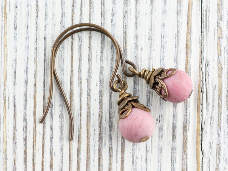 K&ouml;nnte beinhalten: Handgefertigte Ohrringe mit rosa, ovalen Perlen und antiken bronzefarbenen Metallarbeiten. Die Ohrhaken sind ebenfalls bronzefarben. Die Ohrringe werden auf einer wei&szlig;en, verwitterten Holzoberfl&auml;che pr&auml;sentiert.