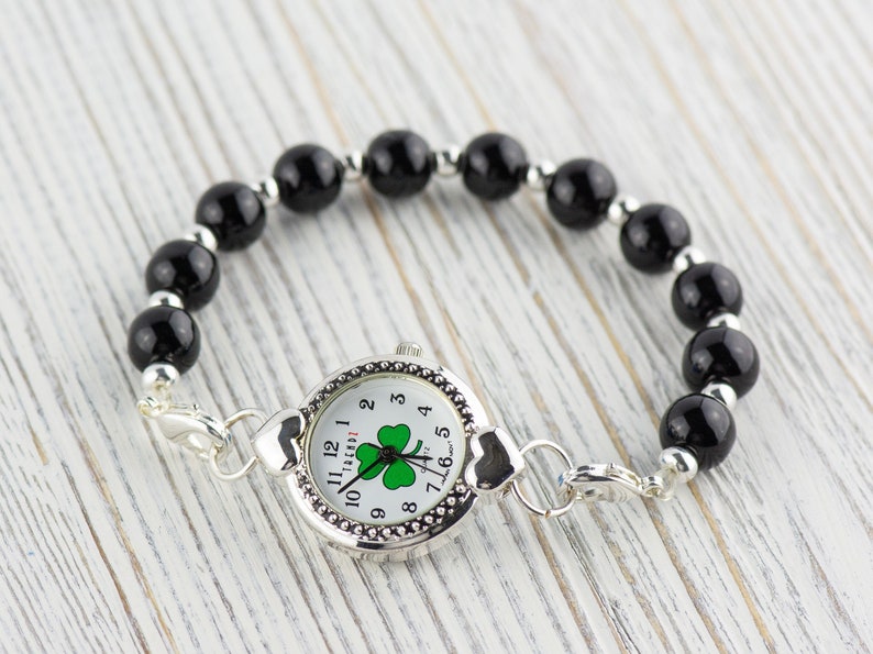 Peut inclure: Un bracelet en perles noires avec un cadran de montre argent&eacute; orn&eacute; d'un tr&egrave;fle &agrave; quatre feuilles. Le cadran de la montre comporte des chiffres romains blancs et un tr&egrave;fle vert au centre.