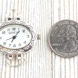 May include: Silver-coloured oval watch face with a white dial and black numerals. The watch face is next to a US quarter for size comparison. The watch reads "GENEVA".