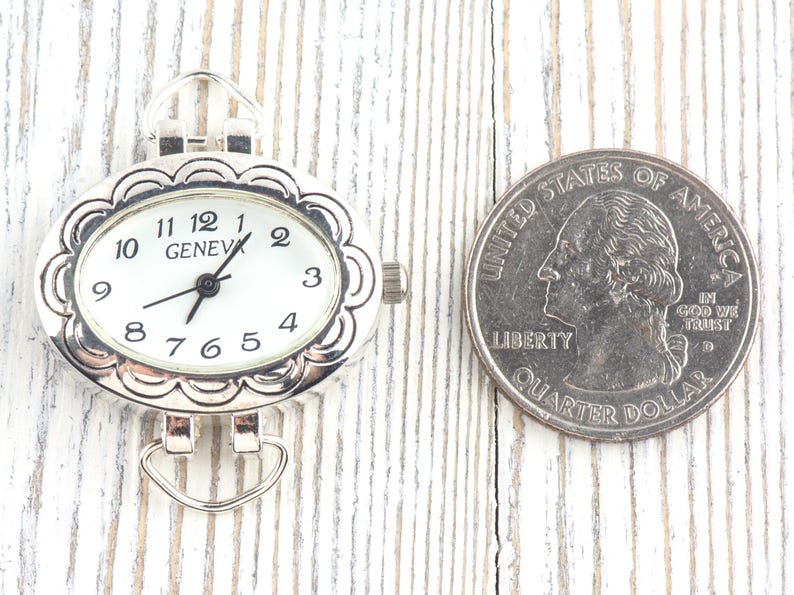 May include: Silver-coloured oval watch face with a white dial and black numerals. The watch face is next to a US quarter for size comparison. The watch reads "GENEVA".