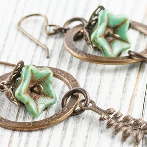 May include: Close-up of two antique bronze earrings. Each earring features a turquoise star-shaped bead, a decorative bronze ring, and a bronze leaf-shaped charm. The earrings are set against a white wooden background.
