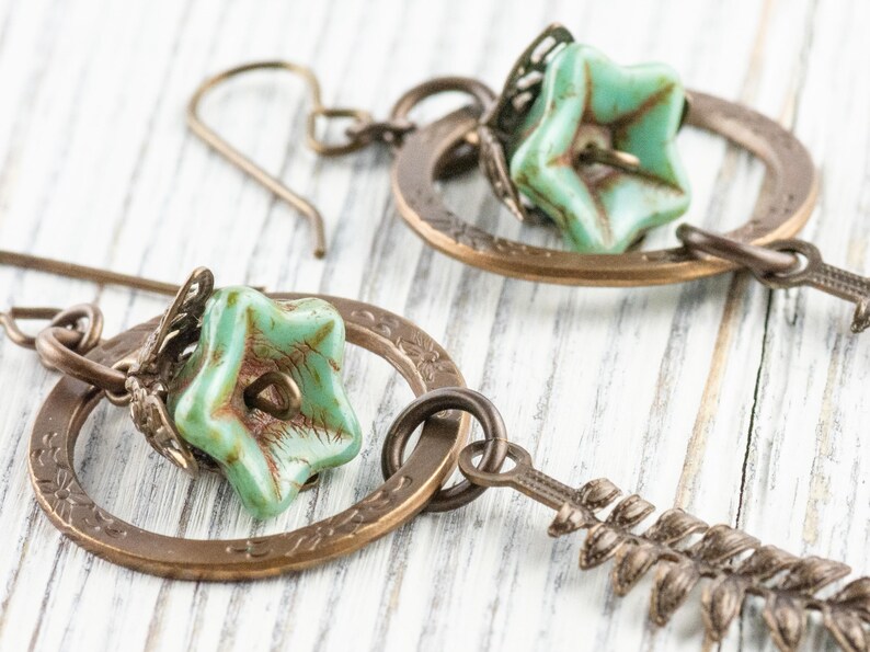 May include: Close-up of two antique bronze earrings. Each earring features a turquoise star-shaped bead, a decorative bronze ring, and a bronze leaf-shaped charm. The earrings are set against a white wooden background.