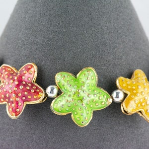 Puede incluir: Un brazalete colorido con cinco abalorios en forma de estrella. Los abalorios son de metal y est&aacute;n esmaltados en rojo, verde, amarillo y azul. Cada abalorio tiene un contorno dorado y est&aacute; decorado con peque&ntilde;os puntos blancos.