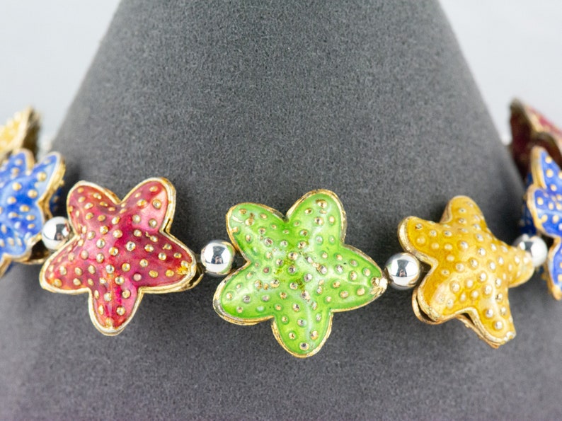 Puede incluir: Un brazalete colorido con cinco abalorios en forma de estrella. Los abalorios son de metal y est&aacute;n esmaltados en rojo, verde, amarillo y azul. Cada abalorio tiene un contorno dorado y est&aacute; decorado con peque&ntilde;os puntos blancos.
