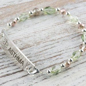 May include: A silver medical alert bracelet with green and pink beads. The bracelet has a tag that reads "ASTHMA" with a medical symbol.