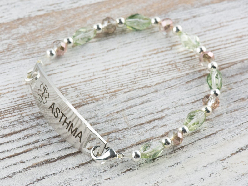 May include: A silver medical alert bracelet with green and pink beads. The bracelet has a tag that reads "ASTHMA" with a medical symbol.