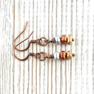 May include: A pair of handcrafted earrings with copper ear wires and decorative beads. Each earring features a silver, bronze, and gold-colored bead arrangement. The earrings are displayed on a white wooden surface.