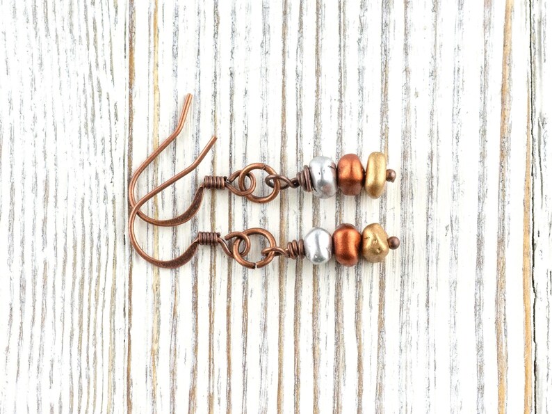 May include: A pair of handcrafted earrings with copper ear wires and decorative beads. Each earring features a silver, bronze, and gold-colored bead arrangement. The earrings are displayed on a white wooden surface.