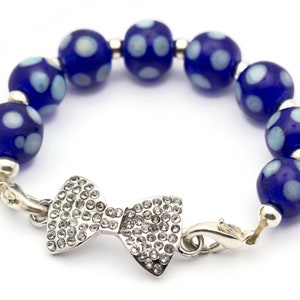 Puede incluir: Un brazalete de cuentas azules y blancas con lunares, con un dije de lazo plateado y un cierre de langosta.
