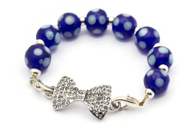 Puede incluir: Un brazalete de cuentas azules y blancas con lunares, con un dije de lazo plateado y un cierre de langosta.