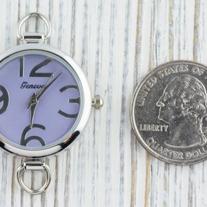 May include: A silver pocket watch with a purple face and black Roman numeral hour markers. The watch has a silver loop on the top and a silver clasp on the bottom.