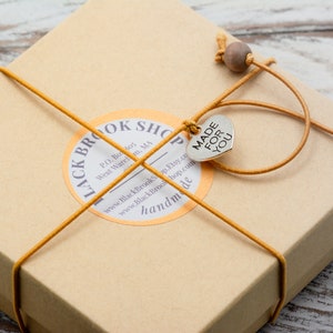 Puede incluir: Una peque&ntilde;a caja de cart&oacute;n marr&oacute;n atada con cuerda amarilla y un dije de coraz&oacute;n plateado que dice "MADE FOR YOU". La caja tiene una etiqueta que dice "Black Brook Shop" e incluye informaci&oacute;n de contacto y la palabra "hecho a mano".