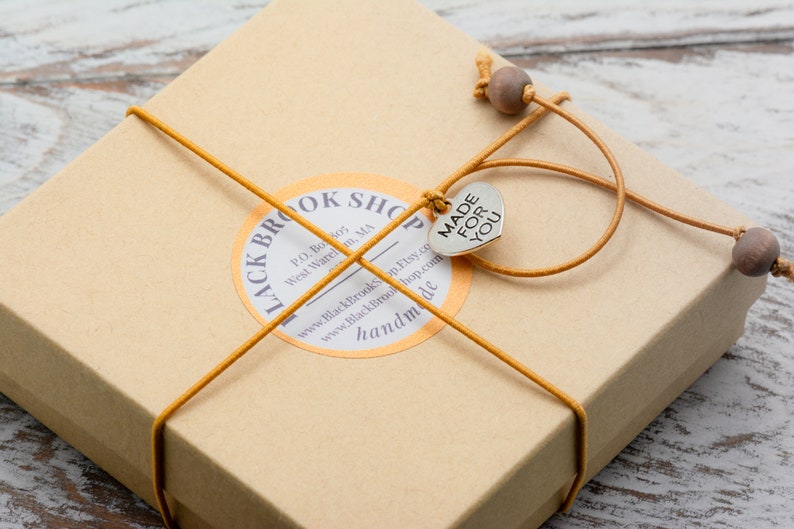 Puede incluir: Una peque&ntilde;a caja de cart&oacute;n marr&oacute;n atada con cuerda amarilla y un dije de coraz&oacute;n plateado que dice "MADE FOR YOU". La caja tiene una etiqueta que dice "Black Brook Shop" e incluye informaci&oacute;n de contacto y la palabra "hecho a mano".